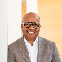 A professional headshot of Bipul Sinha, CEO, Chairman, and Co-Founder of Rubrik, a smiling man of South Asian descent wearing glasses and a light brown blazer over a white shirt.